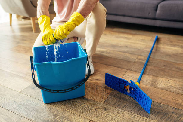Un hombre guapo en acogedora ropa de casa limpia meticulosamente un cubo azul con guantes amarillos. - Foto, imagen