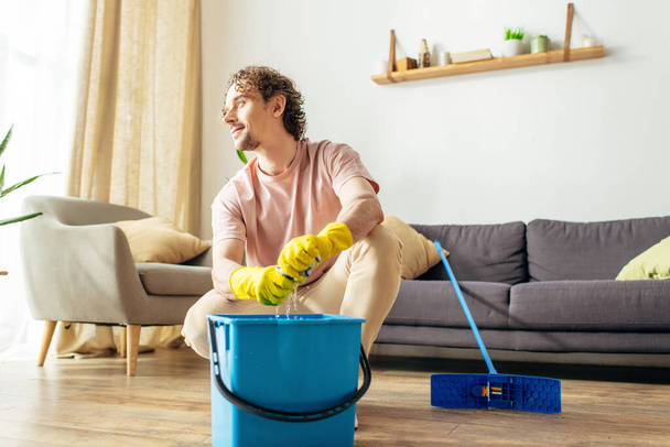 Όμορφος άντρας σε άνετο σπίτι ρούχα καθαρισμού σαλόνι με σφουγγαρίστρα και κουβά. - Φωτογραφία, εικόνα