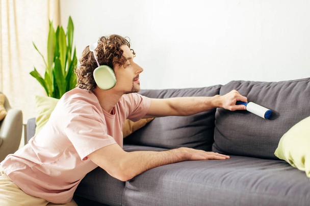 Ένας άντρας με ζεστά ρούχα κάθεται κοντά σε έναν καναπέ, ακούγοντας μουσική με ακουστικά.. - Φωτογραφία, εικόνα