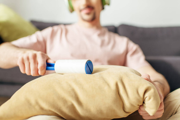 A man in cozy homewear sitting on a couch, holding a sticky roller. - Photo, Image