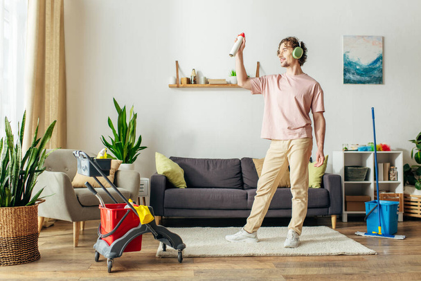 Un hombre en acogedora ropa de casa se encuentra en la sala de estar, sosteniendo limpiador. - Foto, imagen