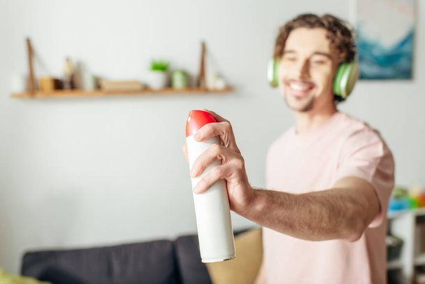 Um homem de roupa de casa aconchegante segura um spray enquanto usa fones de ouvido, absorvido em harmonia musical. - Foto, Imagem