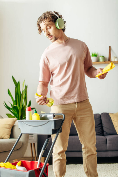 Een man in knusse huiskleding reinigt de woonkamer tijdens het luisteren naar muziek via een koptelefoon. - Foto, afbeelding