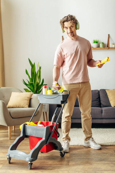 Hezký muž v útulných domácích šatech stojí v obývacím pokoji s vozíkem čistících potřeb. - Fotografie, Obrázek