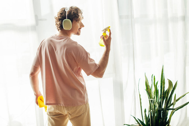Un hombre en ropa de casa acogedora se para frente a una ventana, escuchando música a través de auriculares. - Foto, imagen