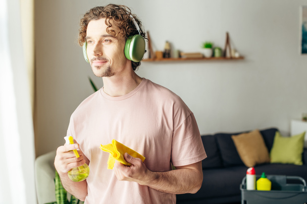 Um homem elegante em roupa de casa acolhedora escuta música através de fones de ouvido enquanto segura um spray. - Foto, Imagem