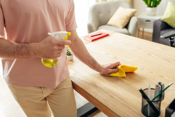 Όμορφος άντρας στο άνετο σπίτι καθαρίζει ένα τραπέζι με ένα κίτρινο πανί. - Φωτογραφία, εικόνα