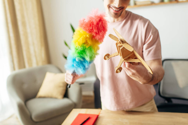 Bonito homem em casa aconchegante segurando um avião de brinquedo e espanador colorido arco-íris. - Foto, Imagem