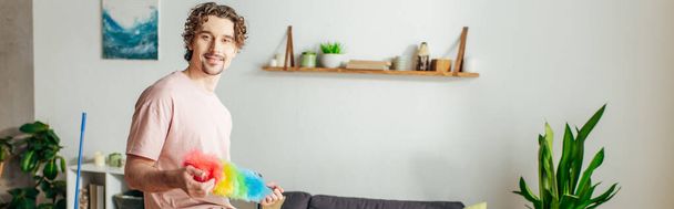 Όμορφος άντρας σε άνετο σπίτι φορώντας ένα πολύχρωμο ξεσκονόπανο σε ένα σαλόνι. - Φωτογραφία, εικόνα