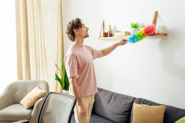Ein gutaussehender Mann in gemütlicher Hauskleidung putzt ein Wohnzimmer mit einem Staubwedel. - Foto, Bild
