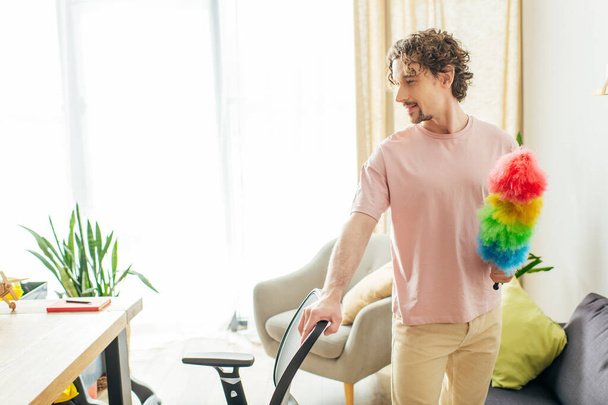 Knappe man in knusse huiskleding met een kleurrijke stofdoek in een woonkamer. - Foto, afbeelding