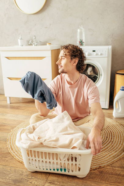 Przystojny mężczyzna w przytulnym homewear siedzi obok kosza na pranie. - Zdjęcie, obraz
