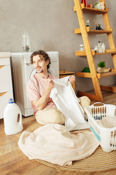 Мужчина в уютной домашней одежде сидит на полу, складывая белую рубашку. - Фото, изображение