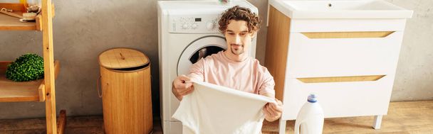 Ev elbiseli bir adam çamaşır makinesinin önünde havlu tutuyor.. - Fotoğraf, Görsel
