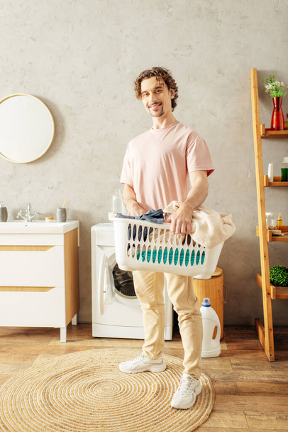 Um homem bonito em roupa de casa aconchegante segurando uma cesta de lavanderia em um quarto. - Foto, Imagem