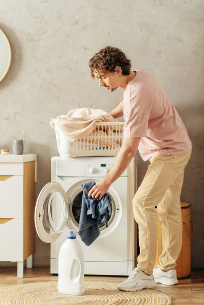 Ένας όμορφος άντρας με ζεστά ρούχα στέκεται δίπλα σε ένα πλυντήριο, έτοιμος να καθαρίσει το σπίτι του.. - Φωτογραφία, εικόνα
