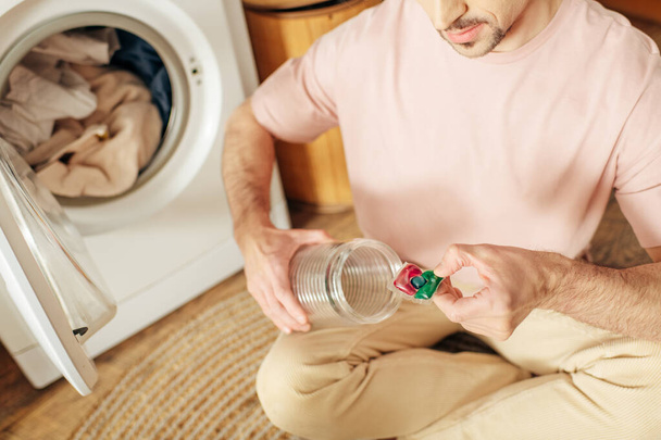 Mężczyzna w przytulnym homewear siedzi na podłodze, trzymając detergent. - Zdjęcie, obraz