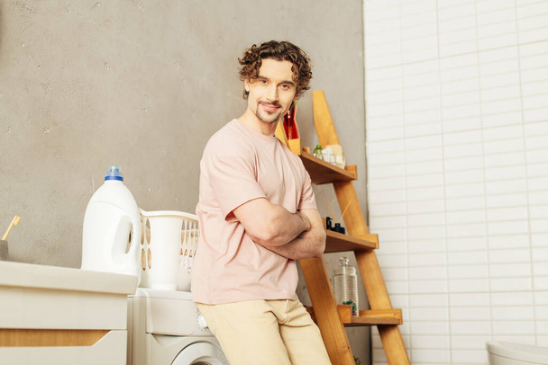 Ein gutaussehender Mann in gemütlicher Homewear steht neben einer Waschmaschine, bereit für den Wäschetag. - Foto, Bild