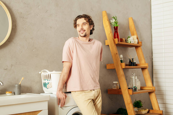 Bonito homem em roupa de casa aconchegante de pé ao lado de uma máquina de lavar roupa em um banheiro. - Foto, Imagem
