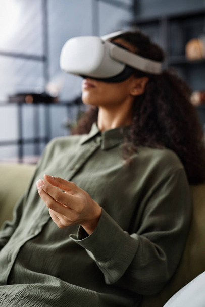 Mise au point sélective plan vertical de la jeune femme portant un casque VR assis sur le canapé dans le salon en utilisant la technologie AR - Photo, image
