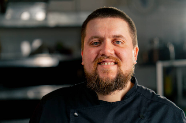 Nahaufnahme in einer professionellen Küche Porträt eines Kochs in schwarzer Jacke in der Küche glückliches Lächeln - Foto, Bild