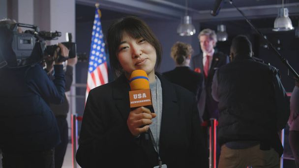 Die asiatische Moderatorin berichtet live aus dem Regierungsgebäude. Journalistin leitet Sendung für politisches Fernsehprogramm von Pressekonferenz mit amerikanischer Politikerin oder US-Präsident - Foto, Bild