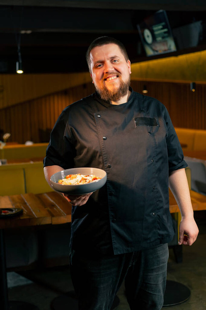 en un restaurante un chef en una chaqueta negra se para con una ensalada preparada sonríe mira a la cámara - Foto, Imagen