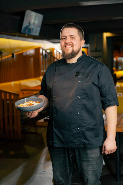 В ресторане шеф-повар в черной куртке стоит с готовым салатом улыбается, смотрит в камеру - Фото, изображение