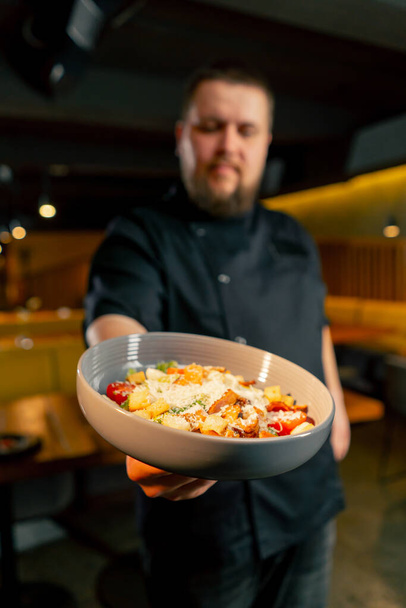 primer plano en un chef de restaurante en una chaqueta negra está sonriendo con una ensalada preparada - Foto, Imagen
