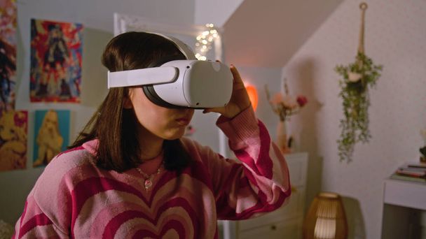 Teenage girl wearing AR headset plays virtual reality online video games in her cozy bedroom. Girl spending leisure time and having fun at home. Concept of modern devices, entertainment and lifestyle. - Photo, Image