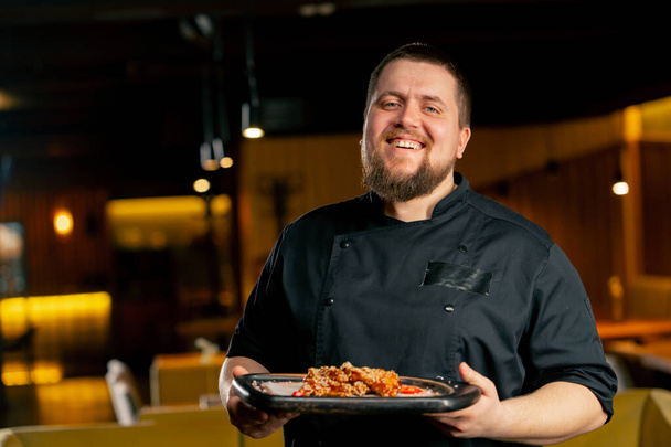 в ресторане шеф-повар в черной куртке стоит улыбаясь с приготовленными куриными крылышками - Фото, изображение