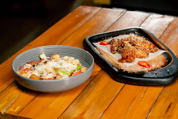 κοντά σε ένα ξύλινο τραπέζι υπάρχει ένα έτοιμο πιάτο σαλάτα Cesar σε ένα γκρι πιάτο και φτερούγες κοτόπουλου - Φωτογραφία, εικόνα