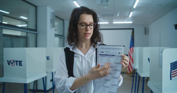 Amerikkalainen nainen puhuu kameran edessä, näyttää paperilehteä, vaatii äänestystä. Kansallinen vaalipäivä Yhdysvalloissa. Äänestyskopit äänestyspaikalla. Kansalaisvelvollisuuden ja isänmaallisuuden käsite. Muotokuva. - Valokuva, kuva