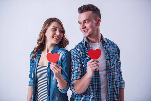 schönes junges Paar in lässiger Kleidung hält zwei rote Papierherzen, schaut einander an und lächelt, isoliert auf weiß - Foto, Bild