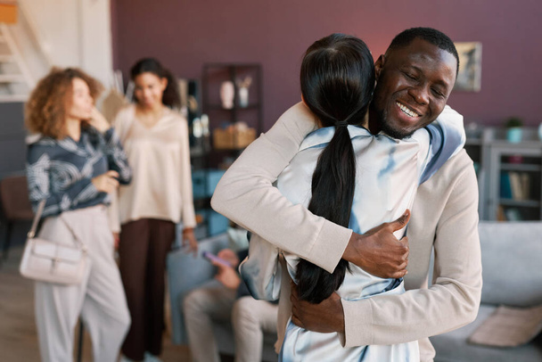 Happy African American guy giving hug to young brunette woman at home party while congratulating her on birthday or some other life event - Photo, Image