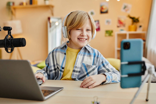 Cute smiling schoolboy in headphones looking at smartphone screen while sitting by desk in front of laptop and watching broadcast - Photo, Image