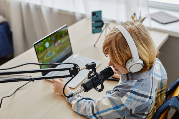 Niño rubio en auriculares hablando en micrófono mientras está sentado en el escritorio delante de la computadora portátil con videojuego en la pantalla y jugando - Foto, imagen