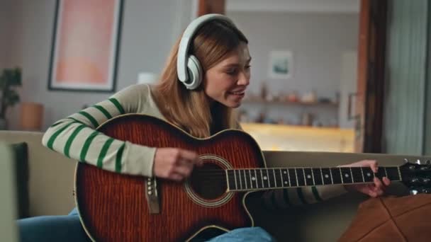 Lány zenész élvezni gitár hang visel fejhallgató otthon kanapén közelről. Mosolygó női gitáros, aki akusztikus hangszeren gyakorolja az akkordokat videóleckét néz a laptopon. Boldog hölgy zenél. - Felvétel, videó