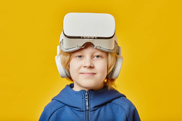 Lindo chico rubio con auriculares VR en la frente mirando a la cámara después de jugar juego virtual mientras está de pie sobre el fondo amarillo - Foto, imagen