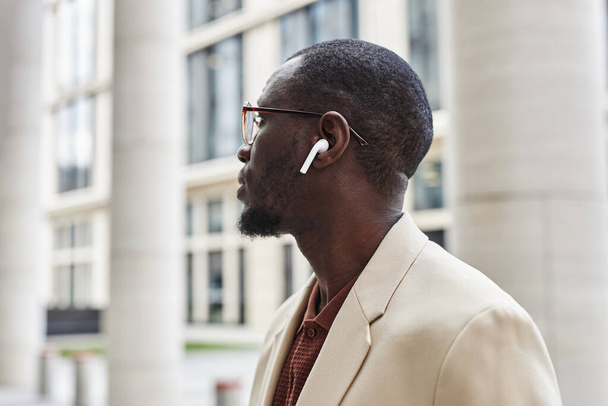 Seitenansicht eines jungen, gelassenen schwarzen Mannes im eleganten weißen Anzug, Brille und Kopfhörer, der Musik hört und etwas Kurioses betrachtet - Foto, Bild