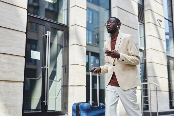 Молодий бізнесмен у формальному одязі та навушники, які шукають вперед під час переїзду в аеропорт вздовж сучасної будівлі та текстових повідомлень у смартфоні - Фото, зображення