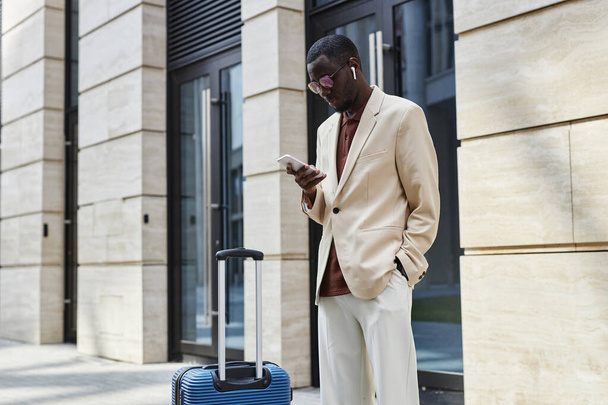 Young serious businessman scrolling or texting in smartphone while standing on sidewalk against modern building and waiting for taxi - Photo, Image