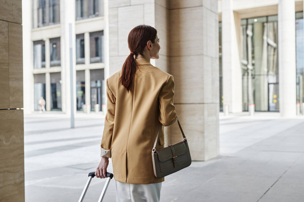 Вид спереду молодої брюнетки бізнес-леді в офіційному одязі тягне валізу під час прогулянки до аеропорту або автовокзалу вздовж сучасної архітектури - Фото, зображення
