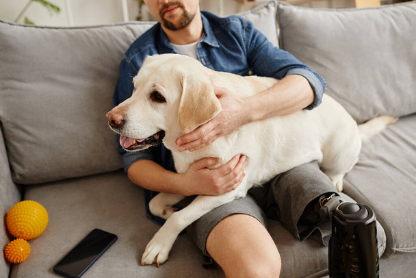 Nahaufnahme eines behinderten Mannes, der einen großen weißen Hund auf dem Schoß hält, während er es sich gemeinsam auf der Couch gemütlich macht - Foto, Bild