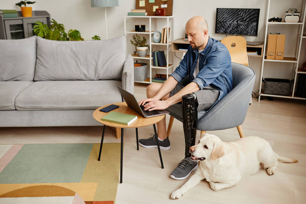 Teljes hosszúságú portré felnőtt férfi műlábbal laptoppal otthon és simogató labrador kutya, másolás helyet - Fotó, kép