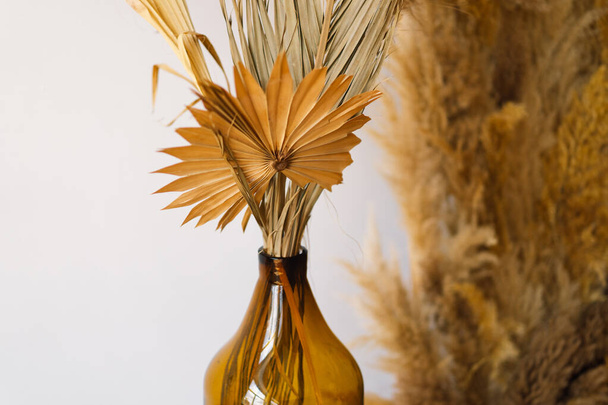 A tasteful arrangement of fluffy pampas grass stalks elegantly placed within an amber glass vase, all set upon a polished wooden stand. The vases warm color complements the neutral tones - Photo, Image
