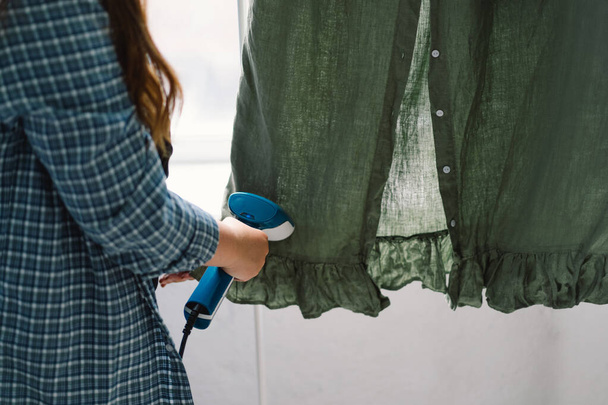 Femme utilisant un vapeur portable bleu sur un vêtement vert à la maison pendant la journée. Lumière naturelle du jour qui éclaire la texture des tissus et la vapeur en action. - Photo, image