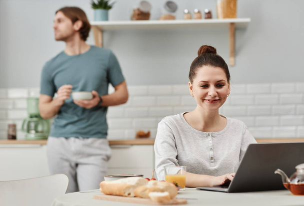 Portrait of smiling young woman using laptop in kitchen during breakfast, copy space - Photo, Image