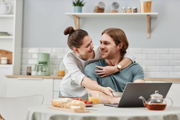Portrait de jeune couple embrassant dans la cuisine avec l'homme utilisant un ordinateur portable et travaillant à la maison, espace de copie - Photo, image