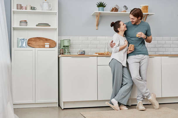 Minimalny portret pełnej długości kochającej się młodej pary obejmującej się w kuchni i uśmiechającej do siebie, przestrzeń kopiująca - Zdjęcie, obraz
